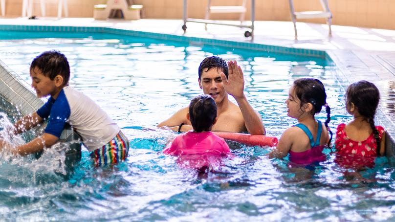 group of kids in pool, one high fives the instructor, another splashes in pool