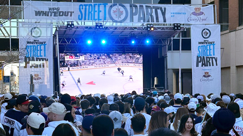 Winnipeg Jets hockey fans standing at the Whiteout Street Party watching a tv showing a NHL playoff game