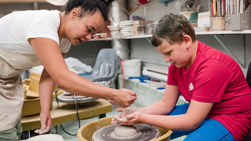 Instructor dropping water onto mound of clay on pottery wheel while student in a red shirt has his hands catching the drips above the clay