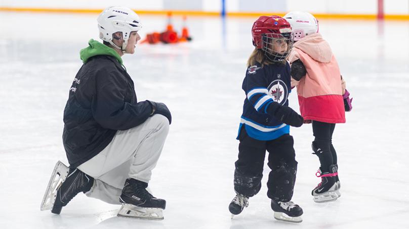 Skating instructor kneeling on ice facing two young skaters during skating lessons