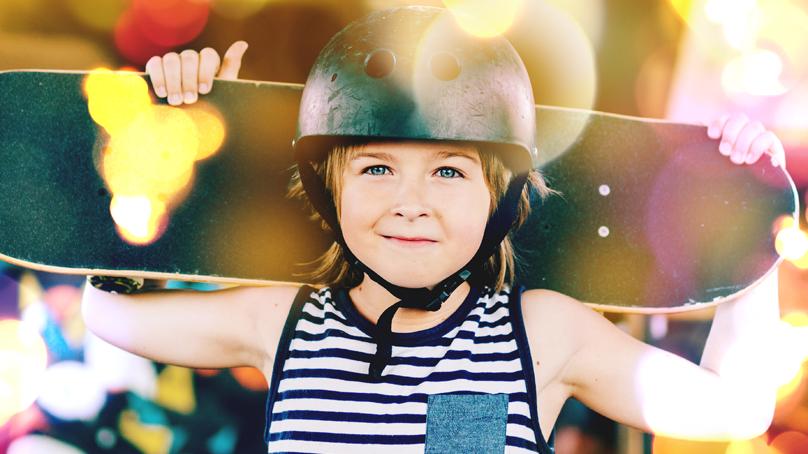 Closeup of skateboarding kid wearing a helmet holding his skateboard up on his shoulders.