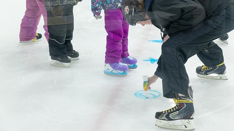 Skating instructor bends down and draws a blue circle on ice next to students