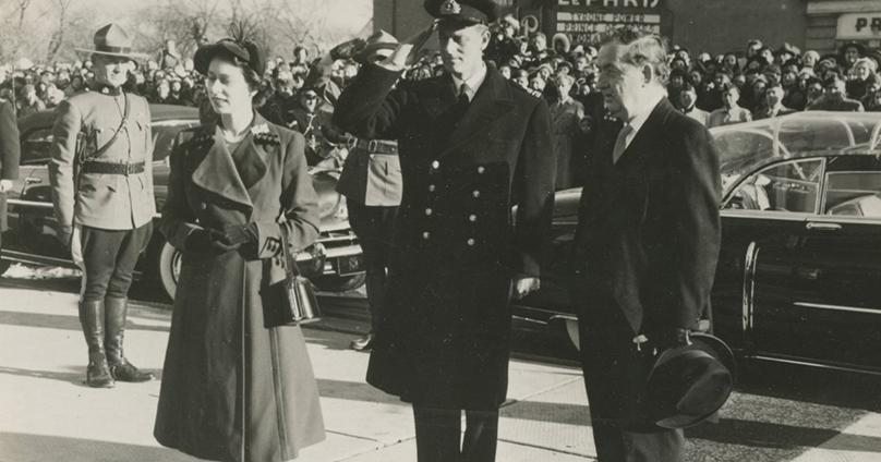 Princess Elizabeth, Duchess of Edinburgh (later Queen Elizabeth II) and the Duke of Edinburgh (or Prince Philip) stand on a sidewalk near the Paris Theatre on Provencher Blvd.